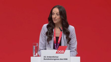 Eine Person steht hinter einem Rednerpult vor einem vollständig roten Hintergrund. Auf dem Pult sind zwei Mikrofone befestigt, links daneben steht ein Glas mit Wasser. Die Person trägt eine helle Jacke über dunkler Kleidung und ein rotes Schlüsselband mit Ausweis um den Hals. Auf der Vorderseite des Rednerpults ist ein rotes Logo mit der Aufschrift „DGB“ sowie darunter der Text „23. Ordentlicher Bundeskongress des DGB“ zu sehen.