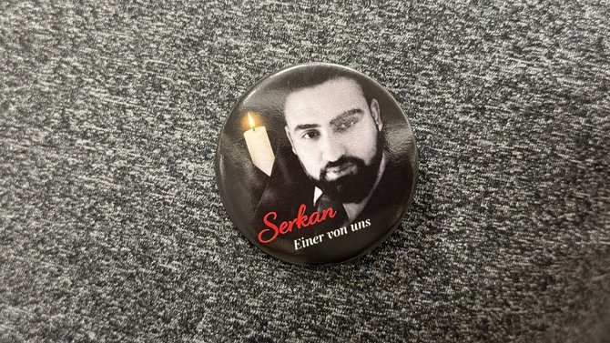 Ein runder Ansteckbutton liegt auf einer grauen, strukturierten Stoffoberfläche. Auf dem Button ist ein schwarz-weißes Porträt zu sehen, daneben eine Kerze. Darunter steht in roter und weißer Schrift der Text: „Serkan – Einer von uns“.