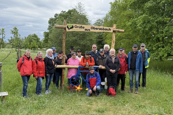 Gruppe von etwa zwanzig Personen steht im Freien auf einer Wiese vor einem hölzernen Torrahmen. Oben am Rahmen befindet sich ein Schild mit der Aufschrift „Grüße aus Oberasbach“ sowie einem grünen Logo und dem Wort „verbindet“. Die Personen tragen überwiegend wetterfeste Jacken, einige Rucksäcke sind sichtbar; eine Person kniet vorne. Am unteren Querbalken ist ein blaues Metallrad mit bunten Blüten befestigt. Im Hintergrund sind Bäume, Sträucher und ein bewölkter Himmel zu sehen.