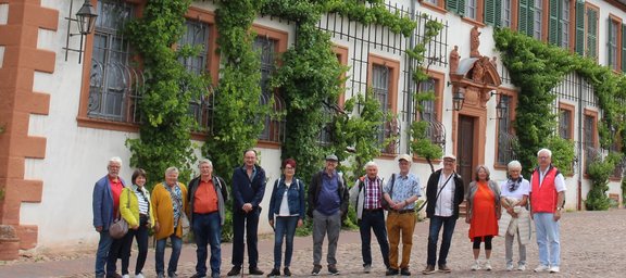 Eine Gruppe von etwa zwölf Personen steht vor einem historischen Gebäude mit vielen Fenstern und grün bewachsenen Rankgittern. Die Personen stehen in einer Reihe auf einem gepflasterten Platz und schauen zur Kamera. Hinter der Gruppe befindet sich eine lange Hausfassade mit hellen Wänden, rot-braunen Rahmen und mehreren Fenstern, die mit Metallgittern versehen sind. An der Fassade wachsen dichte grüne Pflanzen entlang von Spalieren. Es sind mehrere Lampen an der Wand angebracht, und in der Mitte befindet sich ein aufwendig gestalteter Eingang mit einem kleinen Dach und Verzierungen.