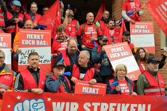 Gruppe von Demonstrierenden auf den Stufen vor einem Backsteingebäude. Viele Personen tragen rote Westen oder rote T‑Shirts mit weißen Aufdrucken, halten rote EVG-Fahnen und blaue Tröten. Mehrere Schilder mit der Überschrift „MERKT EUCH DAS:“ sind sichtbar. Im Vordergrund verläuft ein großes rotes Banner mit der Aufschrift „WIR STREIKEN!“.
