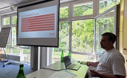 Seminarraum mit großen Fenstern. Eine Person sitzt an einem Tisch mit Laptop vor einer projizierten Präsentationsfolie. Auf der Folie steht die Überschrift „DIE GEFÄHRDUNGSBEURTEILUNG“ mit einer nummerierten Liste. Auf dem Tisch stehen Wasserflaschen und Gläser.