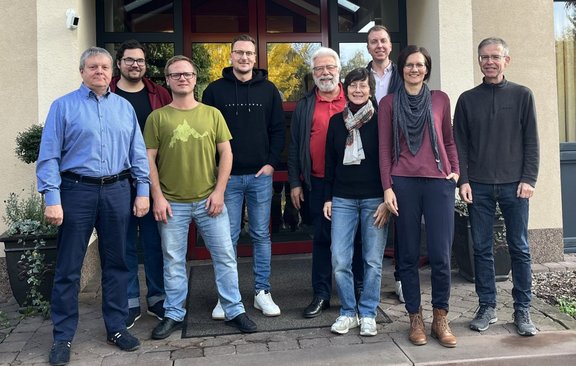 Neun Personen stehen vor dem Eingang eines Gebäudes mit großen Glasflächen und rotem Türrahmen. Die Personen sind in einer Reihe und leicht versetzt angeordnet und tragen überwiegend Freizeitkleidung in Blau-, Grau-, Grün-, Rot- und Schwarztönen. Links und rechts neben dem Eingang stehen Pflanzgefäße. Vor der Gruppe befindet sich ein gepflasterter Bereich mit einer dunklen Fußmatte. Im Hintergrund sind durch die Glasscheiben Reflexionen von Bäumen und Himmel sichtbar.