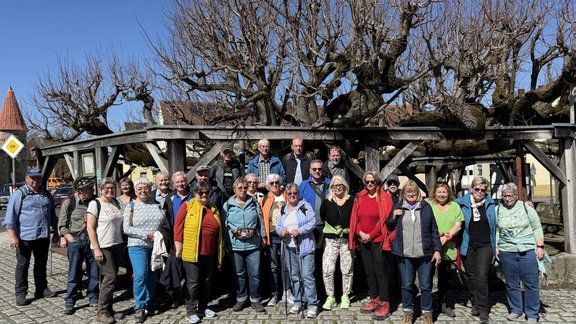 Große Gruppe von etwa zwanzig Personen steht dicht nebeneinander auf einem gepflasterten Platz im Freien. Im Hintergrund befindet sich ein hölzerner Unterstand mit schrägen Balken, darüber stark beschnittene, blattlose Bäume. Dahinter sind Häuser mit hellen Fassaden und roten Ziegeldächern zu sehen, links ein Turm mit spitzem Dach. Die Personen tragen wetterfeste Freizeitkleidung wie Jacken, Westen, Rucksäcke und Wanderschuhe in verschiedenen Farben. Der Himmel ist klar und blau, das Licht wirkt wie an einem sonnigen Tag.