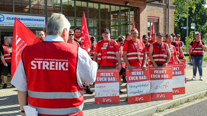 Person mit roter Warnweste, auf deren Rücken „Eisenbahn- und Verkehrsgewerkschaft STREIK!“ steht, spricht vor einer Gruppe von Menschen in roten EVG-Westen und roten T-Shirts. Mehrere rote Fahnen werden gehalten. Vor der Gruppe stehen rote Schilder mit der Aufschrift „MERKT EUCH DAS:“ und handschriftlichen Notizen. Im Hintergrund befindet sich ein Gebäude mit Backsteinfassade und großen Fenstern.
