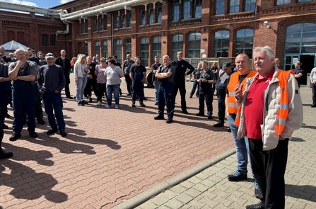 Mehrere Dutzend Personen stehen auf einem gepflasterten Hof vor einem großen Backsteingebäude mit vielen Fenstern und sichtbaren Rohrleitungen an der Fassade. Im Vordergrund stehen zwei Personen mit orangefarbenen Warnwesten, eine davon spricht zu der versammelten Gruppe. Sonniges Wetter mit deutlichen Schatten auf dem Boden.