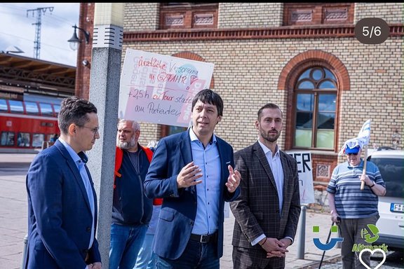 Mehrere Personen stehen vor einem Backsteingebäude und einer Bahnhofsanlage. Im Vordergrund spricht eine Person in dunklem Sakko, während im Hintergrund Demonstrierende Schilder zur medizinischen Versorgung hochhalten. Das Bild zeigt eine Gruppe von Personen auf einem befestigten Platz vor einem Gebäude aus hellen Ziegelsteinen mit Rundbogenfenstern. Links im Hintergrund ist ein roter Zug an einem Bahnsteig zu sehen. Im Vordergrund stehen drei Personen in Anzügen beziehungsweise Sakkos. Die Person in der Mitte spricht mit erhobenen Händen. Hinter der Gruppe stehen weitere Personen mit Protestschildern. Auf einem Schild ist der Text „ärztliche Versorgung in Gefahr“ sowie „+25 h auf Tour statt am Patienten“ zu lesen.