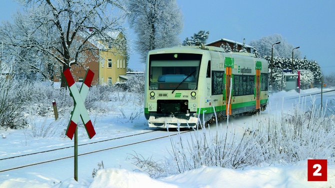 Grün-weißer Triebwagen auf einer schneebedeckten Bahnstrecke. Bäume, Sträucher und Gebäude sind mit Schnee bedeckt. Im Vordergrund steht ein weiß-rotes Andreaskreuz. Unten rechts befindet sich eine rote Markierung mit der Zahl „2“.
