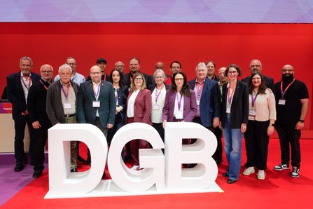 Gruppenfoto mehrerer Personen auf einer roten Bühne. Vor der Gruppe stehen große weiße Buchstaben mit der Aufschrift „DGB“. Alle Personen tragen Namensschilder an Bändern um den Hals. Der Hintergrund ist eine rote Wand ohne weiteren Text.