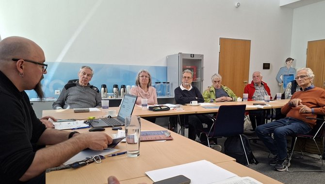 Seitliche Ansicht desselben Besprechungsraums. Mehrere Personen sitzen nebeneinander an einem langen Tisch. Auf dem Tisch stehen Gläser, Tassen, Thermoskannen, Papierunterlagen, ein Laptop und Schreibmaterial. Im Hintergrund sind eine weiße Wand, ein Schrank, eine Tür und eine Wandgrafik mit einer gezeichneten Person zu sehen. Der Raum ist hell beleuchtet, der Boden ist grau, die Tische sind hellholzfarben.