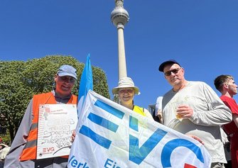 Drei Personen stehen im Freien und halten eine große weiße Fahne mit blauem Schriftzug „EVG“. Eine Person trägt eine orangefarbene Warnweste und hält ein Plakat mit der Aufschrift „Rote Karte dem Kahlschlag!“. Im Hintergrund ist der Berliner Fernsehturm vor blauem Himmel zu sehen.