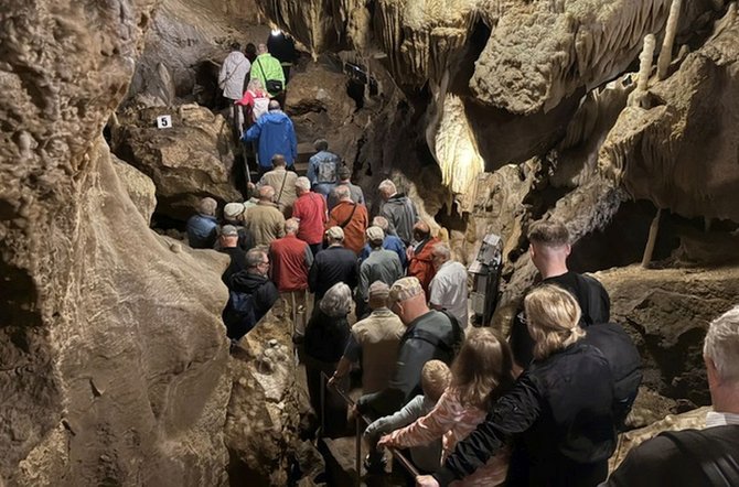 Eine Gruppe von Menschen bewegt sich in einer Tropfsteinhöhle auf einem schmalen Weg mit Geländer nach unten. Die Höhlenwände und Decke sind mit Stalaktiten und Stalagmiten bedeckt und unregelmäßig geformt. Die Szene ist durch künstliches Licht beleuchtet, das die Felsstrukturen hervorhebt. Die Personen sind dicht hintereinander angeordnet und folgen dem Weg, einige halten das Geländer fest.