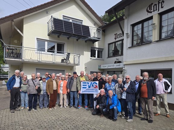 Eine große Gruppe von Personen steht vor einem Gebäude und hält ein Schild mit der Aufschrift „EVG – Eisenbahn- und Verkehrsgewerkschaft“. Etwa 25 bis 30 Personen haben sich in mehreren Reihen vor einem hellen Wohn- oder Gasthausgebäude mit Balkon aufgestellt. Im Zentrum der Gruppe halten zwei Personen ein rechteckiges Schild mit blau-rotem Logo und dem Text „EVG Eisenbahn- und Verkehrsgewerkschaft“. Die Menschen tragen überwiegend Alltagskleidung wie Jacken, Hemden und Hosen; einige haben Brillen oder Mützen. Im Hintergrund ist ein zweistöckiges Gebäude mit cremefarbener Fassade, Fenstern und einem Balkon mit Solarpanel zu sehen. Rechts befindet sich ein Gebäudeteil mit der Aufschrift „Cafe“ sowie einem weiteren Schriftzug über einer Tür. Der Boden besteht aus Pflastersteinen.