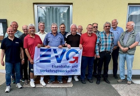 Gruppenfoto von 13 Personen vor einer hellgelben Gebäudewand mit Fenstern. Die Personen stehen in zwei Reihen und halten gemeinsam ein großes weißes Banner mit blau-rotem „EVG“-Logo und dem Text „Eisenbahn- und Verkehrsgewerkschaft“. Die meisten tragen Freizeitkleidung in verschiedenen Farben. Der Boden besteht aus Asphalt und Betonplatten.