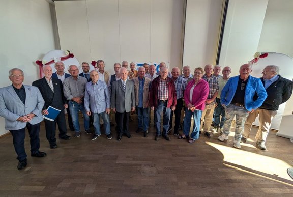 Gruppenfoto mit etwa 20 Personen in einem hellen Innenraum mit Holzboden. Die Personen stehen in mehreren Reihen und blicken zur Kamera. Die Kleidung umfasst Sakkos, Hemden, Jacken und Freizeithosen in verschiedenen Farben. Im Hintergrund sind helle Wände sowie zwei große weiße, bogenförmige Dekorationselemente mit roten Schleifen und Blumen zu sehen. Sonnenlicht fällt von rechts in den Raum und erzeugt helle Lichtflächen und Schatten auf dem Boden.