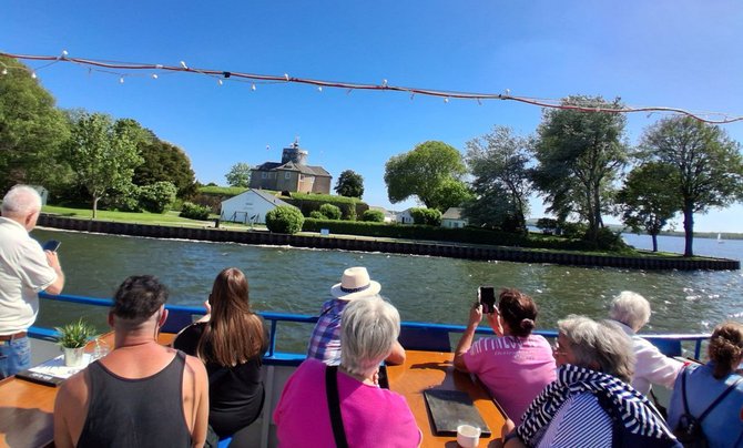 Blick vom Deck eines Schiffes auf mehrere sitzende und stehende Personen, die nach außen auf eine Wasserfläche schauen. Vor ihnen verläuft eine blaue Reling. Am gegenüberliegenden Ufer befindet sich eine kleine Insel oder Uferanlage mit gepflegten Rasenflächen, Bäumen und mehreren Gebäuden, darunter ein größeres Gebäude mit Turm. Einige der Personen halten Smartphones oder Kameras in Richtung Landschaft. Über dem Deck sind gespannte Lichterketten sichtbar.