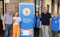 Fünf Personen stehen vor einem Gebäude neben einem Roll‑up‑Banner mit der Aufschrift „Bahnhofsmission“ und „Nächste Hilfe: Bahnhofsmission Osnabrück“. Eine Frau hält einen Scheck in der Hand. Zwei Personen tragen blaue Westen mit einem runden Logo. Im unteren Bereich des Banners sind die Logos von Diakonie und Caritas zu sehen.
