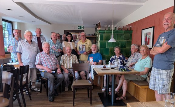 Gruppe von etwa 15 älteren Personen in einem Gastraum mit Holzboden. Einige Personen sitzen an Tischen und auf einer Bank, andere stehen dahinter. Mehrere Personen halten Broschüren oder Hefte in die Kamera. Im Hintergrund stehen ein grüner Kachelofen, Fenster, Wandbilder und ein Notausgangsschild an der Decke. Auf den Tischen stehen Gläser und Flaschen.