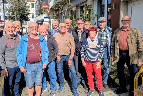 Gruppe von elf Personen steht nebeneinander auf einem Gehweg im Freien. Die Personen tragen Alltagskleidung wie Jacken, Pullover, Hemden, Jeans und Hosen in verschiedenen Farben, darunter Blau, Grau, Braun, Rot und Grün. Eine Person trägt rote Hosen, eine andere eine kurze Hose. Im Hintergrund sind Gebäude mit Schaufenstern, ein Hauseingang, Bäume mit frischen Blättern und Straßenschilder zu sehen. Die Szene ist bei Tageslicht aufgenommen, der Gehweg besteht aus Pflastersteinen.