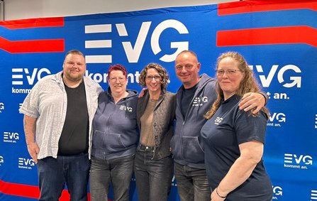 Fünf Personen stehen dicht nebeneinander vor einer blauen Wand mit wiederholtem weißen Schriftzug „EVG kommt an.“ und roten grafischen Elementen. Einige Personen tragen Oberteile mit EVG-Logo.