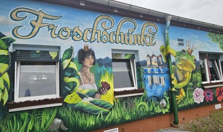 Außenwand eines Gebäudes mit großflächigem Wandgemälde. Sichtbar sind die Aufschrift „Froschänke“, zwei Fenster, Pflanzenmotive, eine gekrönte Froschfigur, eine Personendarstellung, ein Gewässer mit Schlossmotiv sowie Blumen. Unter einem Fenster befindet sich ein Schild mit der Aufschrift „Bitte Vorfahrt“.