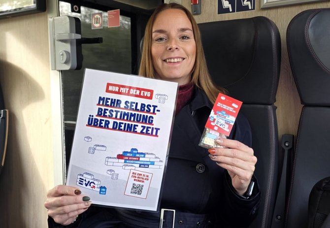 Person sitzt in einem Zug auf einem Sitzplatz und hält einen Flyer mit der Aufschrift „NUR MIT DER EVG MEHR SELBSTBESTIMMUNG ÜBER DEINE ZEIT“ sowie eine kleine rote Verpackung. Auf dem Flyer sind zusätzlich die Texte „BIS ZUM 31.10. EVG-MITGLIED WERDEN!“ und ein QR-Code sichtbar. Im Hintergrund sind die Innenausstattung des Zuges und Hinweisschilder zu sehen.