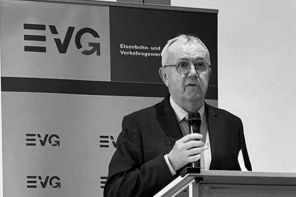 Eine Person steht an einem Rednerpult und spricht in ein Handmikrofon vor einem Banner mit dem EVG-Logo. Das Foto ist in Schwarz-Weiß gehalten. Die Person trägt ein Sakko, Hemd und Krawatte und steht hinter einem Pult. In einer Hand wird ein Mikrofon gehalten. Die Aufnahme zeigt die Person von der Brust aufwärts.