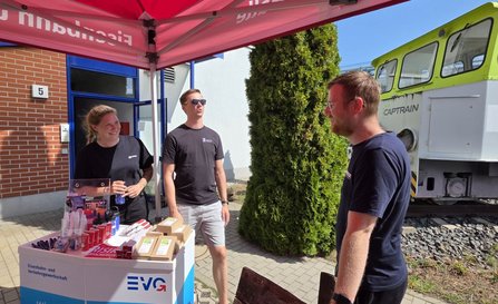 Unter einem roten Pavillon mit EVG-Aufdruck stehen drei Personen an einem Infostand. Auf dem Tresen liegen Werbeartikel, Stifte, Trinkflaschen und mehrere kleine Schachteln. Rechts im Hintergrund steht eine grün-weiße Lokomotive mit der Aufschrift „CAPTRAIN“ auf einem kurzen Gleisabschnitt. Daneben sind ein Gebäude, Bäume und befestigte Wege zu sehen.