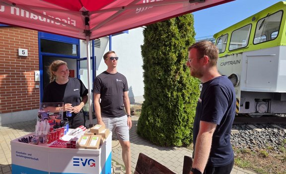 Unter einem roten Pavillon mit EVG-Aufdruck stehen drei Personen an einem Infostand. Auf dem Tresen liegen Werbeartikel, Stifte, Trinkflaschen und mehrere kleine Schachteln. Rechts im Hintergrund steht eine grün-weiße Lokomotive mit der Aufschrift „CAPTRAIN“ auf einem kurzen Gleisabschnitt. Daneben sind ein Gebäude, Bäume und befestigte Wege zu sehen.