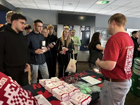 Innenraum mit Informationsstand. Auf einem Tisch liegen gefaltete weiße Textilien, Schlüsselbänder und Flyer. Hinter dem Tisch steht eine Person in rotem Shirt mit großem QR-Code-Aufdruck auf dem Rücken und spricht mit mehreren Personen vor dem Stand. Im Hintergrund befinden sich Automaten, beleuchtete Anzeigetafeln und weitere Personen.