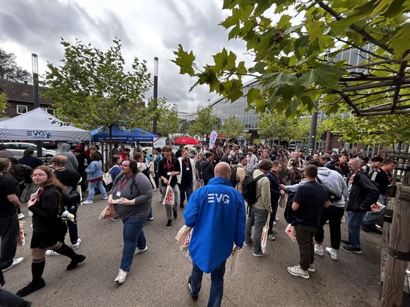 Außenbereich auf einem Platz mit Bäumen und mehreren Informationsständen. Links steht ein weißer Pavillon mit der Aufschrift „EVG JUGEND“, daneben ein blauer Pavillon. Viele Personen bewegen sich über den Platz oder stehen in Gruppen zusammen. Mehrere Personen tragen helle Stofftaschen mit rotem Aufdruck und Namensschilder an roten Bändern. Im Vordergrund steht eine Person in einer blauen Jacke mit großem „EVG“-Schriftzug auf dem Rücken. Der Himmel ist stark bewölkt.
