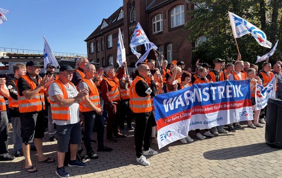 Eine große Gruppe von Personen steht im Freien vor einem Backsteingebäude und trägt überwiegend orangefarbene Warnwesten. Mehrere Personen halten weiße Fahnen mit blau-roten Aufdrucken. Im Vordergrund ist ein großes Banner mit der Aufschrift „WARNSTREIK“ zu sehen; darunter steht „Für faire Löhne und Gute Arbeit für alle Beschäftigten!“. Auf dem Banner sind außerdem EVG-Logos sichtbar. Links im Hintergrund verläuft eine erhöhte Gleisanlage. Das Bild ist bei sonnigem Wetter mit blauem Himmel aufgenommen.