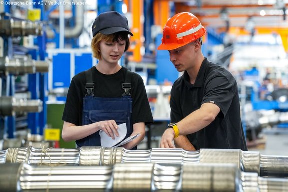 Zwei Personen stehen in einer industriellen Produktionshalle nebeneinander. Eine Person trägt dunkle Arbeitskleidung und eine Mütze und hält ein Notizpapier in den Händen. Die andere Person trägt ein schwarzes Shirt und einen orangefarbenen Schutzhelm und zeigt mit einer Hand auf ein metallisches Bauteil im Vordergrund. Im Vordergrund liegen mehrere parallel angeordnete, glänzende Metallrohre. Im Hintergrund sind Maschinen, Regale und weitere industrielle Ausstattung zu sehen.