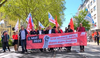 Demonstrationszug auf einer breiten Straße mit Straßenbahnschienen. Mehrere Personen gehen nebeneinander und tragen ein großes rotes Banner mit der Aufschrift „1. Mai 2026 – Erst unsere Jobs, dann eure Profite“ sowie einem erhobenen Faustymbol und dem Kürzel „DGB“. Hinter und neben dem Banner werden rote Fahnen sowie weiße Fahnen mit blauem „EVG“-Schriftzug getragen, darunter auch Fahnen mit dem Zusatz „Jugend“. Die Straße ist von Bäumen mit grünem Laub gesäumt, links stehen Wohngebäude mit gelber Fassade, rechts weitere Gebäude. Der Himmel ist blau, Tageslicht.