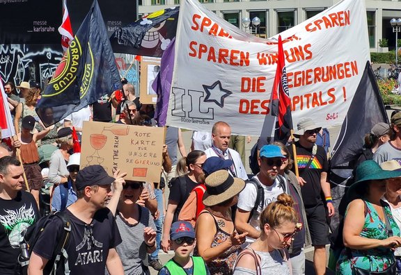 Menschenmenge bei einer Demonstration auf einem Platz in Karlsruhe. Mehrere Fahnen und Transparente werden getragen. Ein großes weißes Banner mit roter Schrift enthält mehrfach das Wort „SPAREN“ sowie den Text „SPAREN WIR UNS DIE REGIERUNGEN DES KAPITALS!“. Ein Pappschild zeigt die Aufschrift „ROTE KARTE FÜR UNSERE REGIERUNG“ und „TAX THE RICH“. Im Hintergrund sind Gebäude mit der Beschriftung „messe Karlsruhe“ zu sehen.