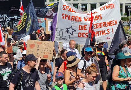 Menschenmenge bei einer Demonstration auf einem Platz in Karlsruhe. Mehrere Fahnen und Transparente werden getragen. Ein großes weißes Banner mit roter Schrift enthält mehrfach das Wort „SPAREN“ sowie den Text „SPAREN WIR UNS DIE REGIERUNGEN DES KAPITALS!“. Ein Pappschild zeigt die Aufschrift „ROTE KARTE FÜR UNSERE REGIERUNG“ und „TAX THE RICH“. Im Hintergrund sind Gebäude mit der Beschriftung „messe Karlsruhe“ zu sehen.