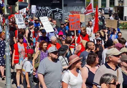 Demonstrationszug mit vielen Teilnehmenden auf einer Straße. Zahlreiche rote Fahnen und handgemalte Schilder sind sichtbar. Lesbare Aufschriften umfassen unter anderem „STELLENABBAU stoppen“, „Geld für … statt für Krieg & Aufrüstung“, „Medikamente retten Gesundheit! Unbezahlbar!“ und „Ihre Profite unsere Krise“. Im Hintergrund befinden sich Fassaden mit Graffiti.