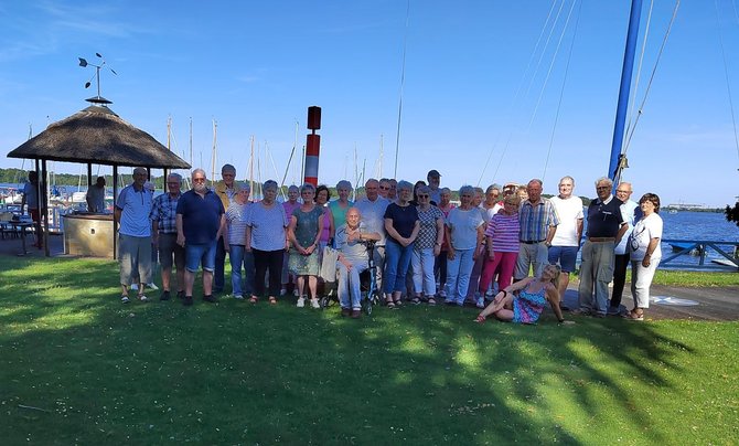 Etwa 30 Personen stehen auf einer Rasenfläche für ein Gruppenfoto. Im Hintergrund befinden sich ein Hafen mit zahlreichen Segelbootmasten, Wasser und ein rundes Pavillongebäude mit Reetdach. Rechts ist der Mast eines Segelboots sichtbar. Der Himmel ist klar und blau.