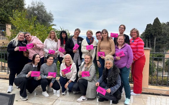 Eine Gruppe von 17 Personen steht und sitzt im Freien vor einer niedrigen Metallbrüstung. Alle halten ein pinkfarbenes Blatt Papier mit dunkler Aufschrift in den Händen. Die Personen sind in zwei Reihen angeordnet: eine vordere Reihe sitzend oder hockend, eine hintere Reihe stehend. Im Hintergrund sind Bäume, Sträucher und Gebäude zu sehen. Der Himmel ist bedeckt.