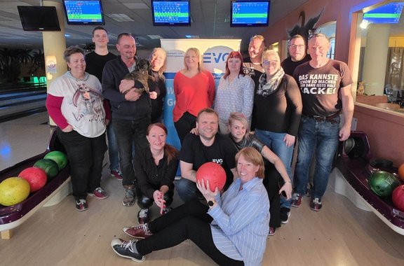 Gruppe von Erwachsenen in einer Bowlinghalle, stehend und sitzend auf der Bahn, umgeben von Bowlingkugeln in Regalen; über ihnen hängen Anzeigetafeln, im Hintergrund stehen Bowlingbahnen und ein Banner mit der Aufschrift „EVG“, eine Person hält eine Bowlingkugel, eine andere ein kleines Tier; Holzboden und bunte Kugeln prägen die Szene.