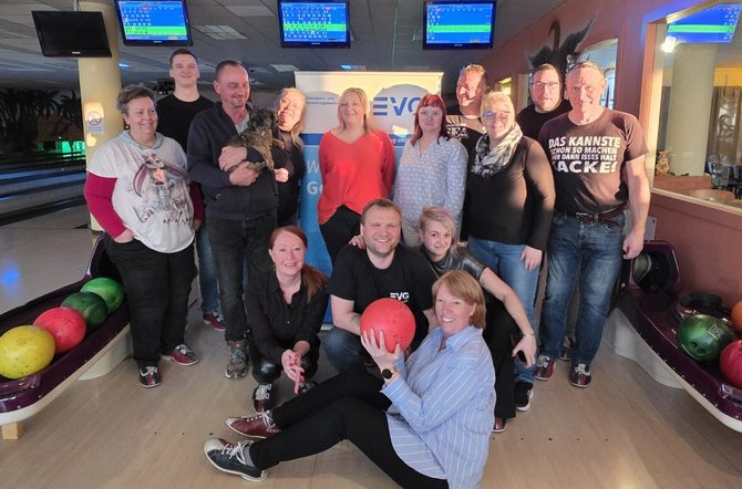 Gruppe von Erwachsenen in einer Bowlinghalle, stehend und sitzend auf der Bahn, umgeben von Bowlingkugeln in Regalen; über ihnen hängen Anzeigetafeln, im Hintergrund stehen Bowlingbahnen und ein Banner mit der Aufschrift „EVG“, eine Person hält eine Bowlingkugel, eine andere ein kleines Tier; Holzboden und bunte Kugeln prägen die Szene.