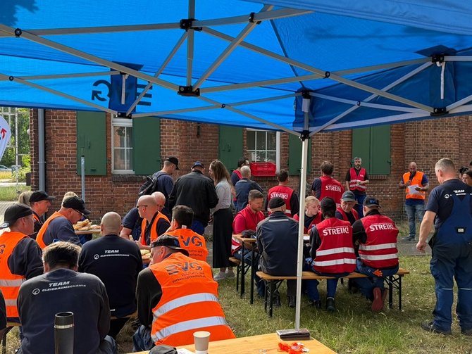 Unter einem blauen EVG‑Zelt sitzen mehrere Personen an Tischen, einige tragen Westen mit „STREIK!“ oder „EVG“. Die Szene spielt draußen vor einem Backsteingebäude mit Fenstern und grünen Fensterläden. Über den Personen steht ein blaues Pavillonzelt, auf dessen Stoff „EVG“ zu lesen ist. Darunter und daneben sitzen mehrere Personen an Biertischen und Bänken. Einige tragen orange Warnwesten mit EVG‑Aufdruck, andere rote Westen mit großer weißer Aufschrift „STREIK!“. Auf den Tischen stehen Becher, Flaschen und kleine Gegenstände. Im Hintergrund stehen weitere Personen, darunter auch eine Person mit Warnweste vor der Gebäudewand. Der Boden ist eine Mischung aus Gras und befestigtem Untergrund. Links und rechts sind weitere Gruppen von sitzenden und stehenden Personen zu sehen.