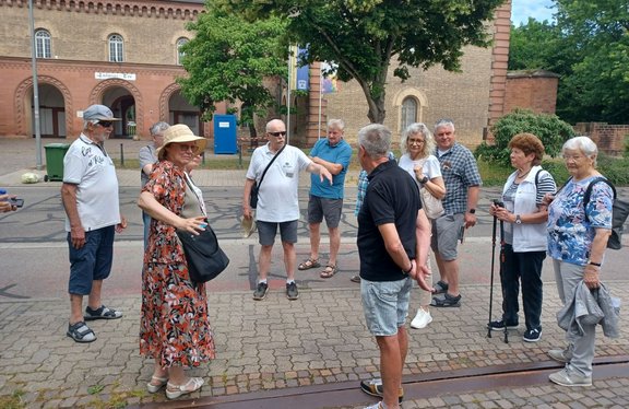 Eine Gruppe von etwa zehn erwachsenen Personen steht auf einem gepflasterten Gehweg vor einem Backsteingebäude mit Rundbögen. In der Mitte gestikuliert eine Frau mit Hut und gemustertem Kleid, rechts daneben steht ein Mann mit dem Rücken zur Kamera. Mehrere Personen tragen sommerliche Kleidung, einige haben Sonnenbrillen, Rucksäcke oder Gehstöcke. Im Hintergrund sind Bäume, eine blaue mobile Toilette und eine Mülltonne zu sehen.