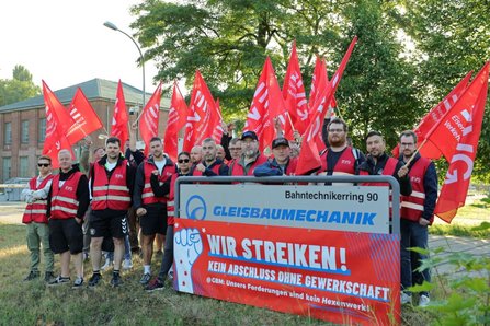 Gruppe von Personen in roten Warnwesten versammelt sich hinter einem Schild mit den Aufschriften „Bahntechnikerring 90“ und „GLEISBAUMECHANIK“. Zahlreiche rote EVG-Fahnen sind über der Gruppe zu sehen. Auf dem davor angebrachten Banner steht „WIR STREIKEN! KEIN ABSCHLUSS OHNE GEWERKSCHAFT“. Die Szene befindet sich im Freien mit Bäumen und einem Backsteingebäude im Hintergrund.