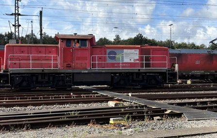 Rote Rangierlokomotive auf einem Gleisfeld mit mehreren parallel verlaufenden Bahngleisen und Oberleitungen. In einem Seitenfenster der Lok ist eine Person sichtbar. Auf der Lok befindet sich ein Plakat mit Text und Grafikelementen.