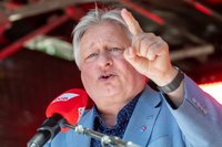 Eine Person steht hinter mehreren Mikrofonen mit roten Windschützen, auf denen „DGB“ steht. Die Person trägt ein hellblaues Sakko und ein dunkelblaues Hemd. Eine Hand ist erhoben, der Zeigefinger zeigt nach oben. Im Hintergrund sind dunkle Strukturen und rote Lichtakzente sichtbar.