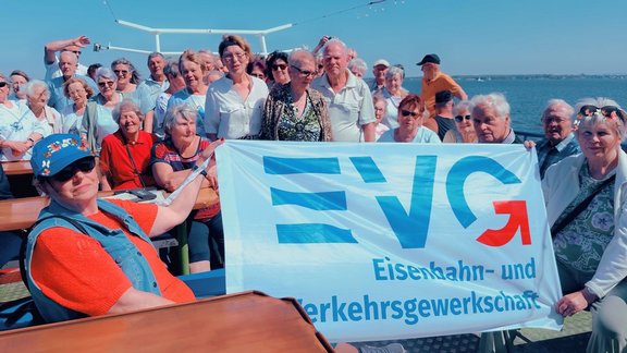 Große Gruppe von vielen Personen steht dicht zusammen auf dem Deck eines Schiffes unter blauem Himmel. Im Vordergrund hält eine Person sitzend oder angelehnt ein großes, weißes Banner mit blauem Schriftzug „EVG“ und darunter „Eisenbahn- und Verkehrsgewerkschaft“; rechts am Banner ist ein roter Pfeil nach oben dargestellt. Die Personen tragen überwiegend helle Kleidung; einige tragen Brillen oder Kopfbedeckungen. Im Hintergrund sind Reling, Aufbauten des Schiffes und Wasser zu erkennen.