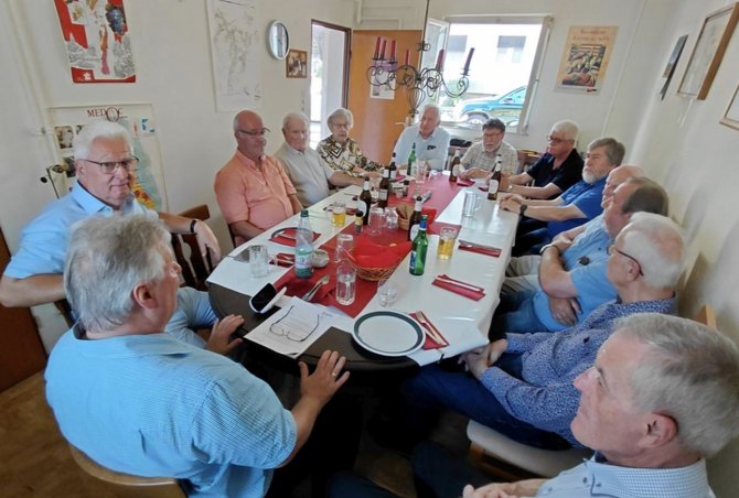Rund ein Dutzend Personen sitzen an einem länglichen Esstisch in einem hellen Raum. Der Tisch ist mit weißer Tischdecke, rotem Tischläufer, Tellern, Gläsern, Flaschen und Servietten gedeckt. An den Wänden hängen Karten, Bilder und ein Spiegel. Eine offene Tür und ein Fenster befinden sich im Hintergrund.