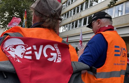 Zwei Personen mit orangefarbenen EVG-Westen stehen mit dem Rücken zur Kamera. Auf einem roten Rucksack ist ein weißes Motiv mit der Aufschrift „EVG REBELLION“ und einer dargestellten Person mit Schutzhelm abgebildet. Im Hintergrund sind Fahnen, weitere Teilnehmende und ein mehrstöckiges Gebäude mit Fensterreihen zu sehen.