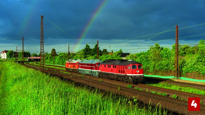 Eine rote Diesellokomotive fährt auf Bahngleisen durch eine grüne Landschaft. Über der Strecke stehen Oberleitungsmasten, im Hintergrund spannt sich ein Regenbogen vor dunklen Wolken über den Himmel. Unten rechts befindet sich eine rote Markierung mit der Zahl „4“.